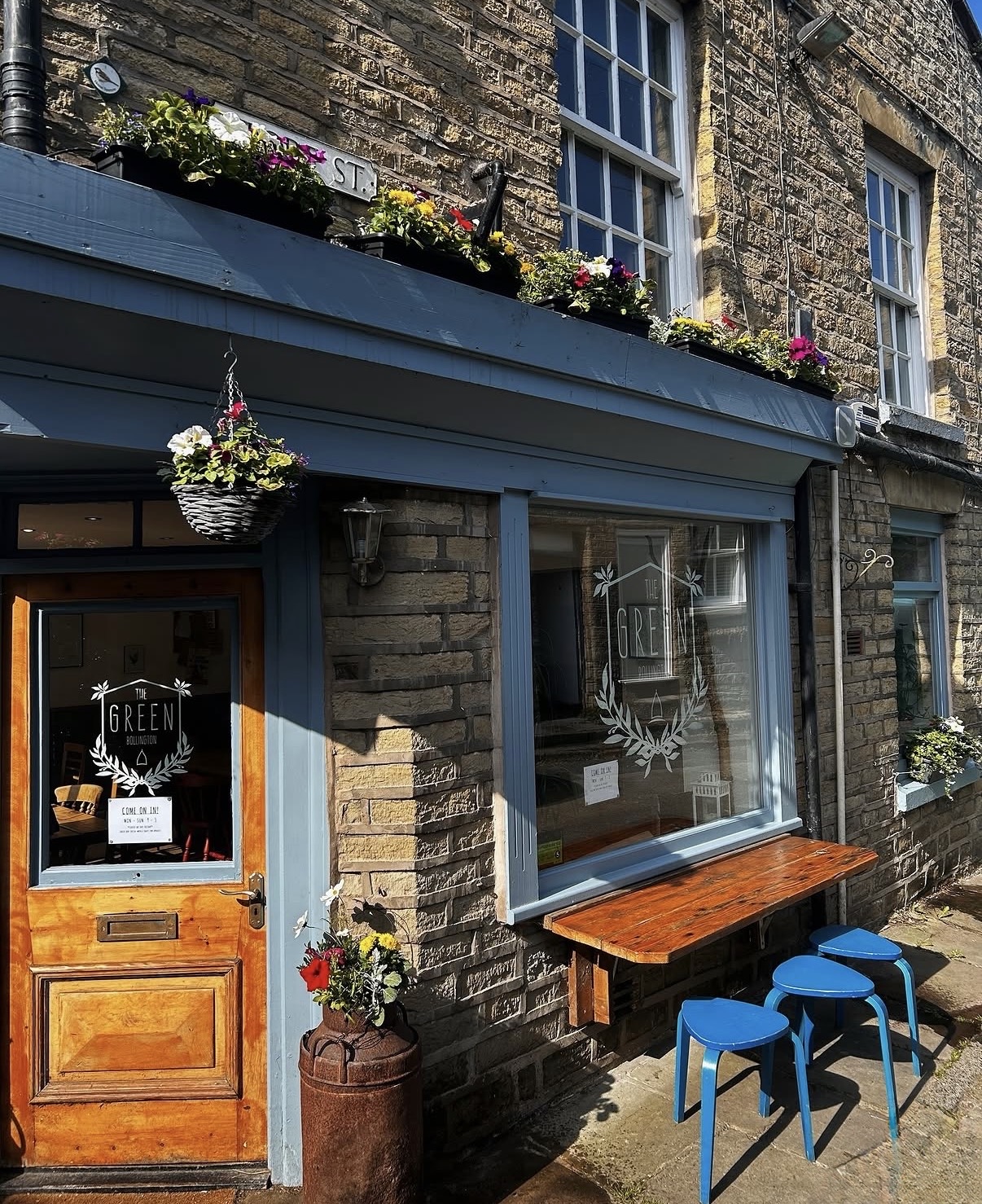 The Green's wooden front door and window with the café logo, a hanging basket, and a window bench with blue stools